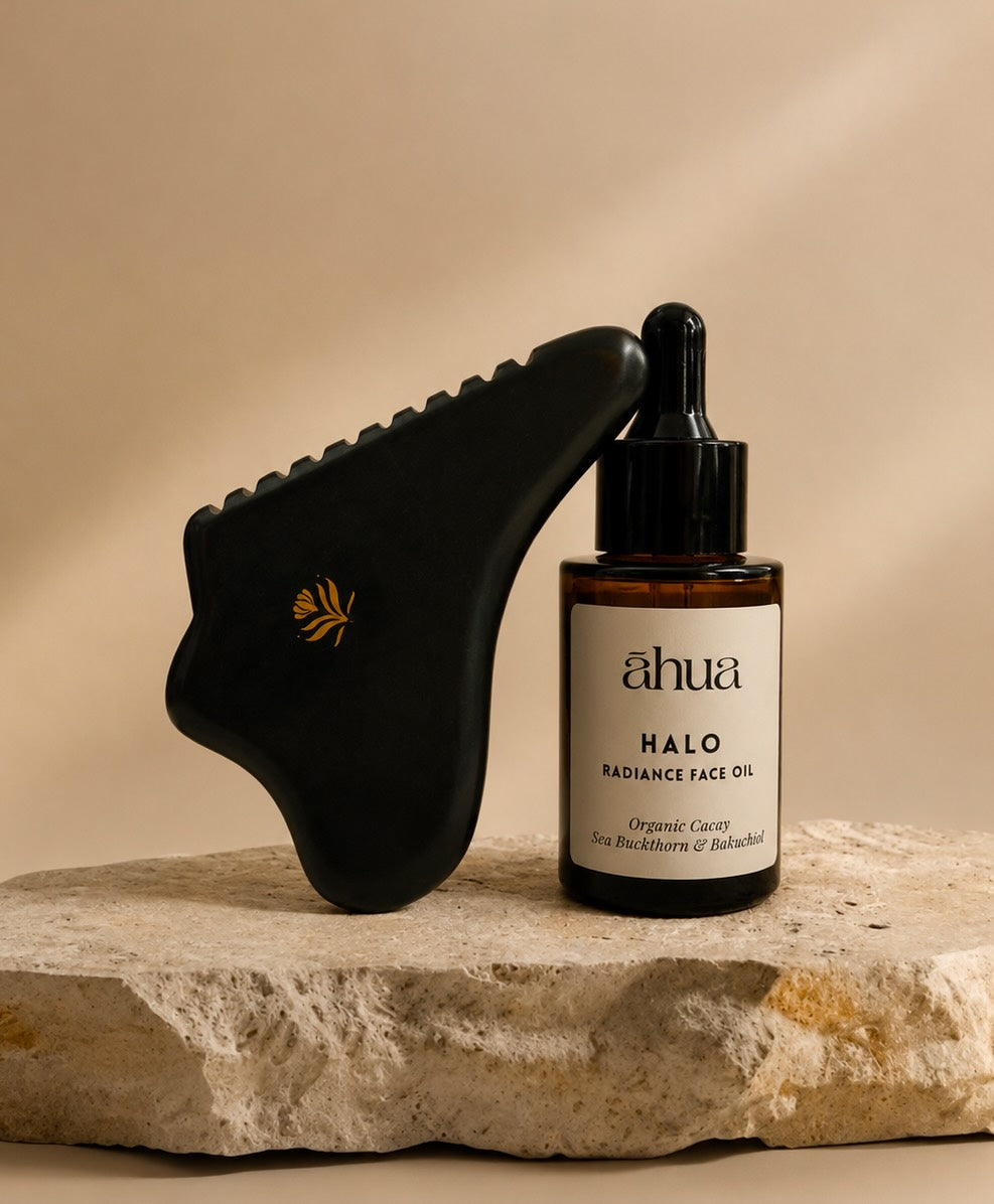 A black gua sha massage tool with a small gold leaf emblem leans against an amber glass dropper bottle labeled “ahua HALO RADIANCE FACE OIL” on a textured stone slab against a warm beige background.
