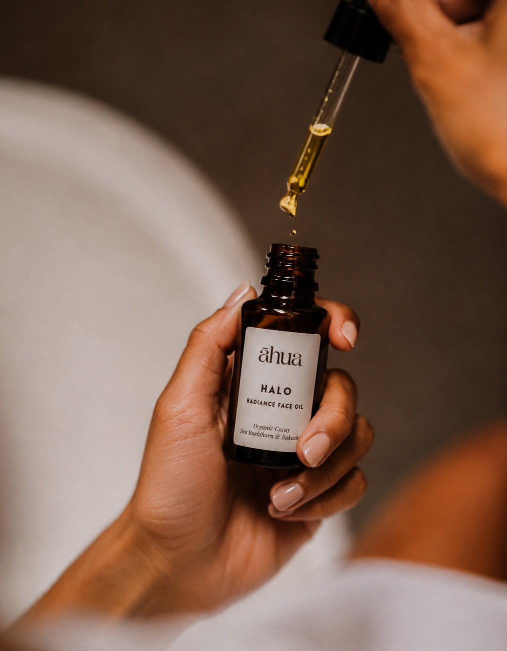 A close-up of a hand holding an amber glass bottle of ahua Halo Radiance Face Oil while another hand drops the golden oil from a glass dropper above the open bottle.