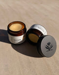 An amber glass jar of Restore Deep Repair Balm with its white label visible, alongside a dark gray metal lid embossed with a botanical logo, set on a textured beige surface under direct sunlight.
