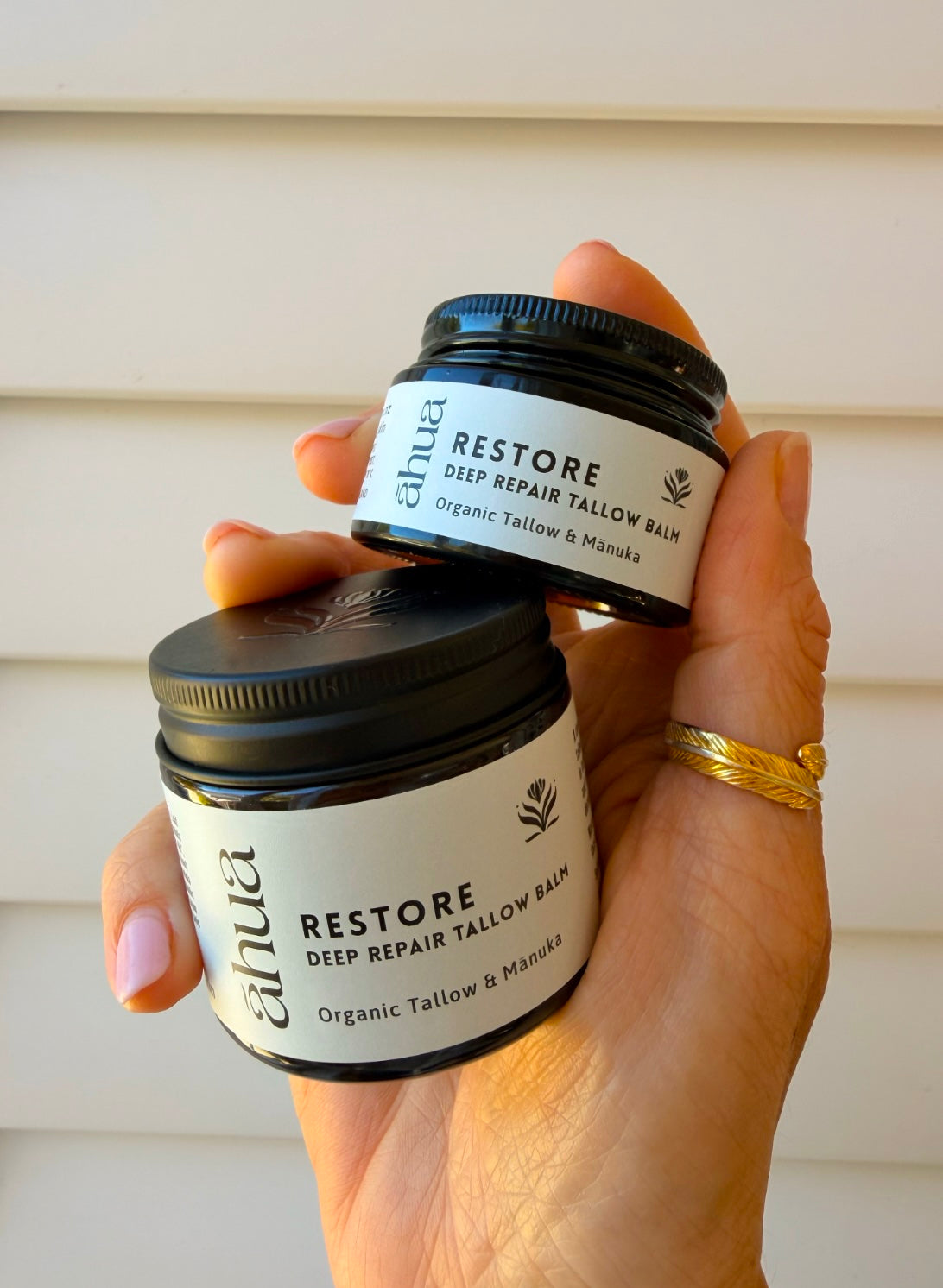A hand holds two small jars of Ahua Restore Deep Repair Tallow Balm against a light beige siding background. The hand is wearing a gold ring.