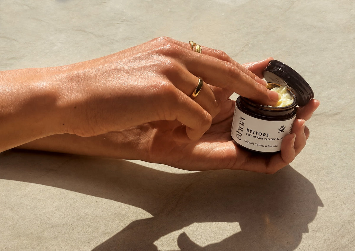 A close-up of a person's hands holding an open dark jar of skincare cream and applying it. The jar has a white label with the brand name 'AHUA' and product name 'RESTORE'.