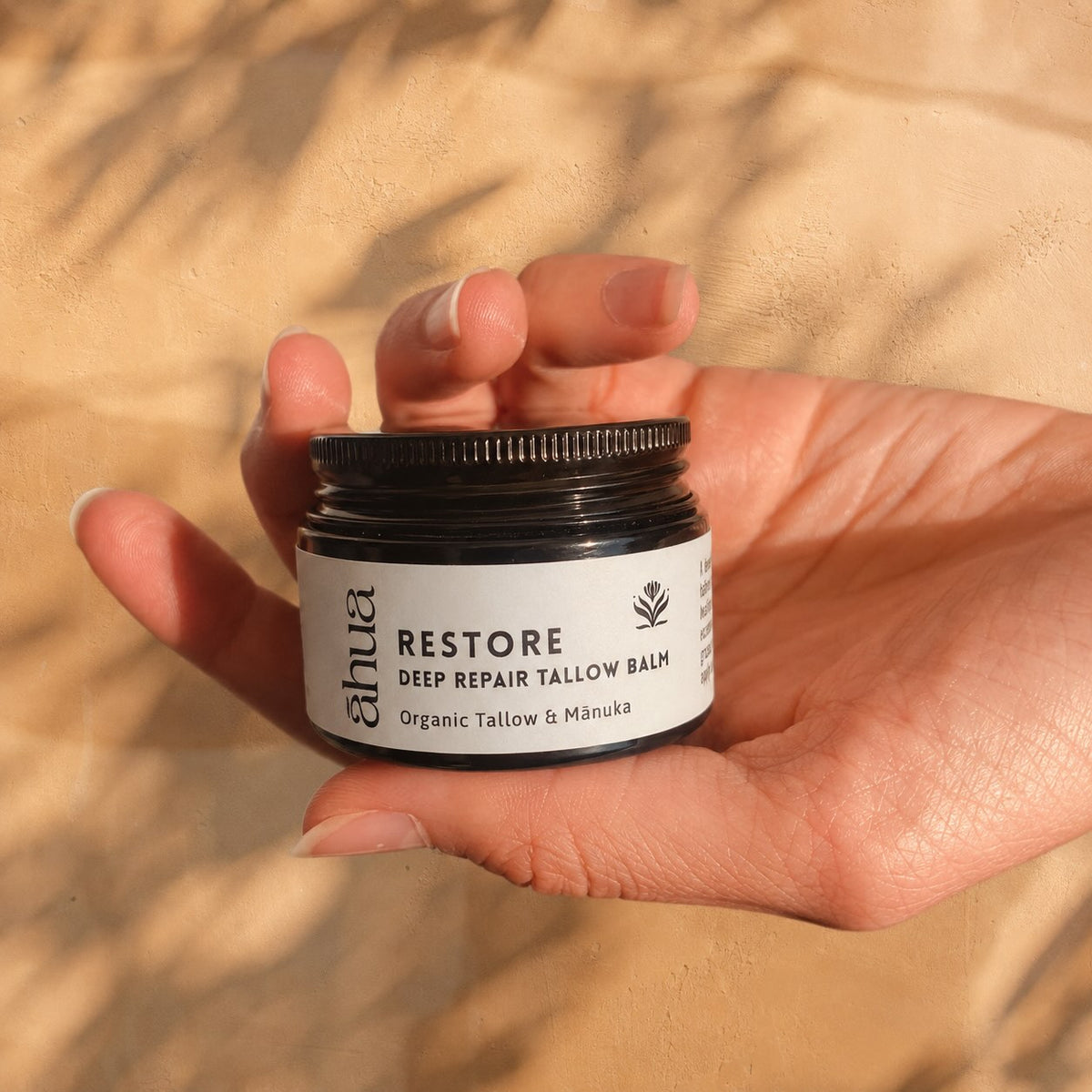 A hand holding a small black jar of Ahua Restore Deep Repair Tallow Balm against a sandy beige background with soft leaf shadows.
