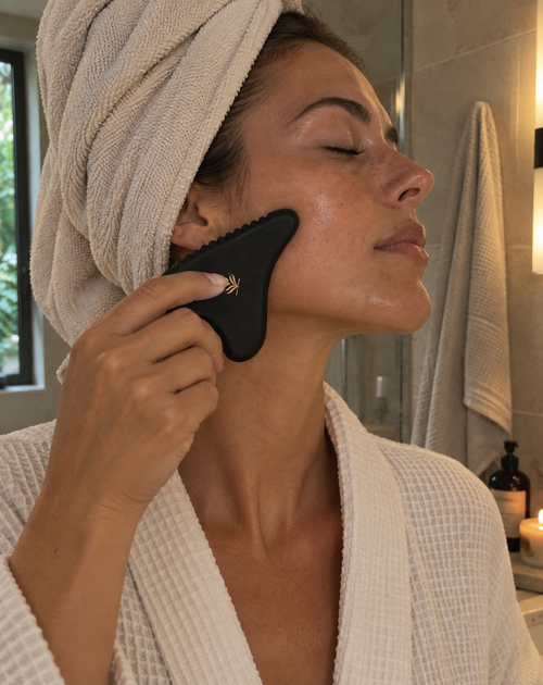 A woman in a white robe with a beige towel wrapped around her head uses a black facial gua sha massage tool on her face in a bathroom setting.